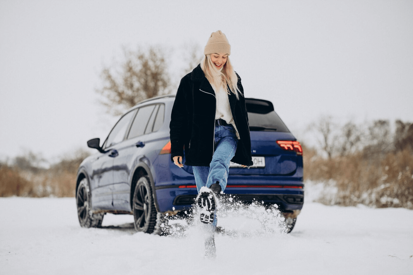 Image of a smiling woman kicking up snow with a parked suv in the snow.
