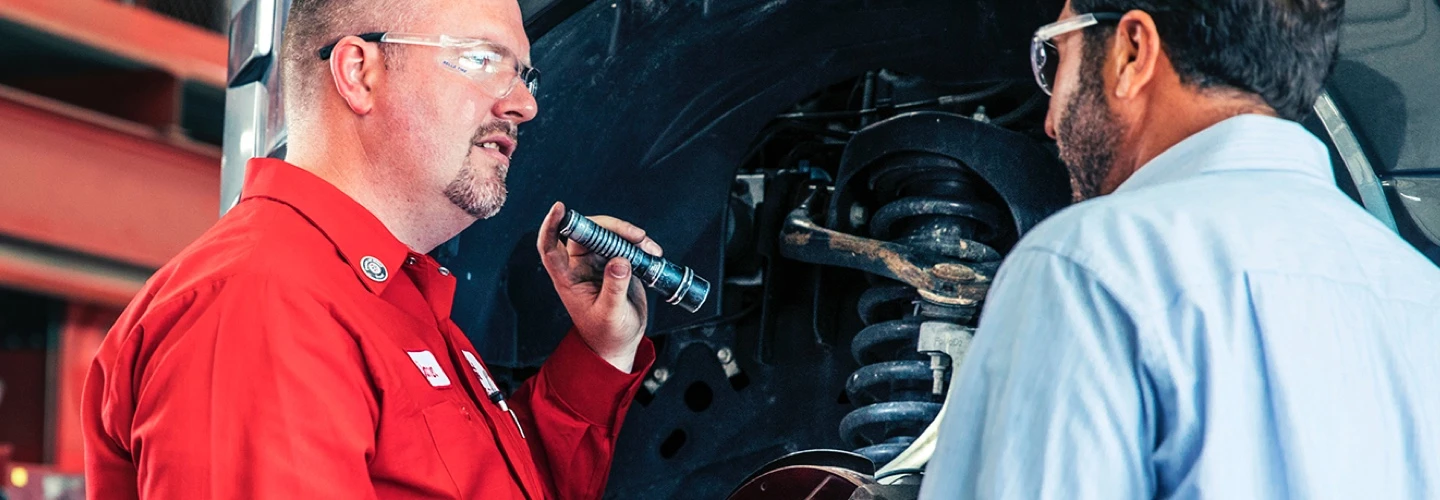 A Tire World Auto Repair tech in a red shirt and wearing safety glasses is inspecting struts with a flashlight. He’s speaking with a Tire World Auto Repair customer.